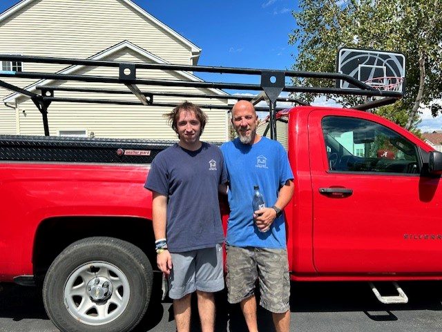 Brian and his helper standing proudly next to their red service truck loaded with tools and ladders, ready to work.